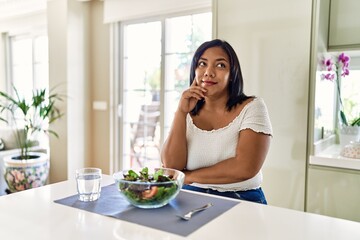 Obraz na płótnie Canvas Young hispanic woman eating healthy salad at home with hand on chin thinking about question, pensive expression. smiling with thoughtful face. doubt concept.