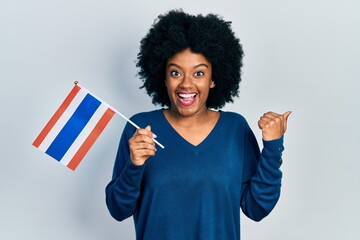 Young african american woman holding thailand flag pointing thumb up to the side smiling happy with open mouth