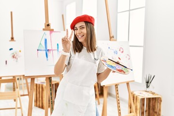 Young artist woman painting on a canvas at art studio smiling looking to the camera showing fingers...