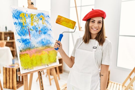 Young hispanic artist woman wearing french beret drawing using paint roller at art studio.