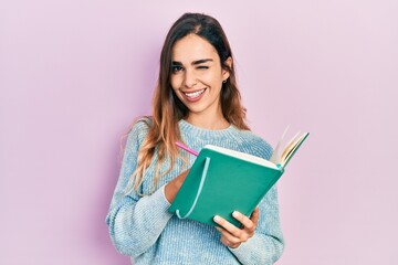 Young hispanic girl reading and writing book winking looking at the camera with sexy expression, cheerful and happy face.