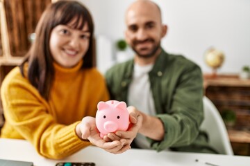 Young hispanic couple smiling happy holding piggy bank sitting on the table at home.