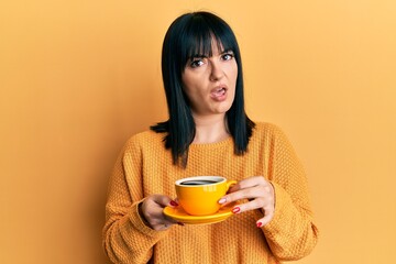 Young hispanic woman holding coffee in shock face, looking skeptical and sarcastic, surprised with open mouth