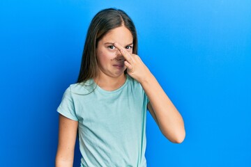 Beautiful brunette little girl wearing casual white t shirt smelling something stinky and disgusting, intolerable smell, holding breath with fingers on nose. bad smell