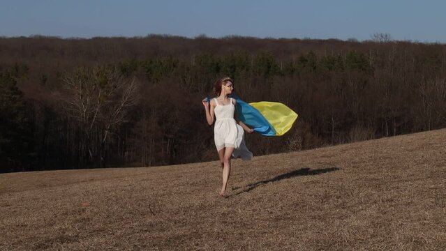 Woman running with wrapped ukrainian yellow blue flag flutters waving in the wind. National symbol of Ukraine. Patriotic lady with makeup. Stand with Ukraine, support and solidarity
