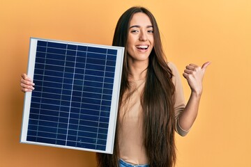 Young hispanic girl holding photovoltaic solar panel pointing thumb up to the side smiling happy...