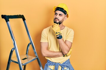 Handsome man with beard by construction stairs wearing hardhat with hand on chin thinking about question, pensive expression. smiling with thoughtful face. doubt concept.