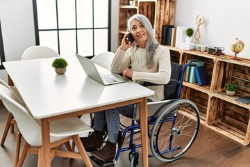 Middle age grey-haired woman talking on the smartphone sitting on wheelchair at home