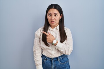 Young latin woman standing over blue background pointing aside worried and nervous with forefinger,...