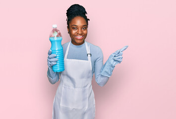 Young african american woman wearing apron holding detergent bottle smiling happy pointing with hand and finger to the side