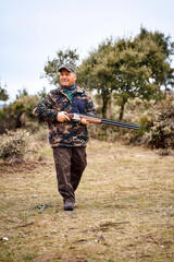 Full body aged male hunter in camouflage outerwear and cap carrying gun and looking away while...