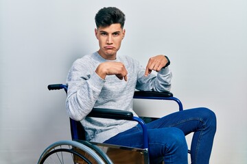 Young hispanic man sitting on wheelchair pointing down looking sad and upset, indicating direction...