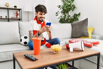 Young hispanic hooligan man using smartphone supporting soccer team at home