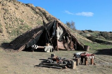 A reed tent on a mountain slope. There are old furniture and fire in front of the tent.