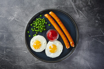 Classic breakfast of fried eggs, sausages and green peas on dish on gray background