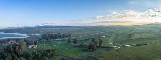 Aerial view of a green grassland, trees and a mountain covered with snow in background at sunrise, Golan Heights, Israel.