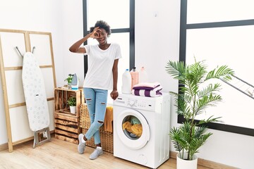 African young woman doing laundry at home smiling happy doing ok sign with hand on eye looking through fingers