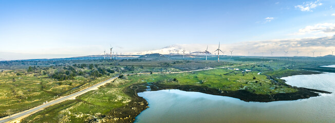Panoramic aerial view of wind turbine farm in a grassland, a lake and mountain covered with snow in background, Golan Heights, Israel.