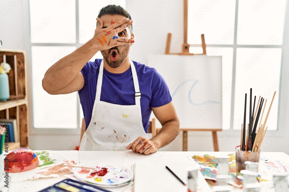 Sticker Young hispanic man with beard at art studio with painted face peeking in shock covering face and eyes with hand, looking through fingers with embarrassed expression.