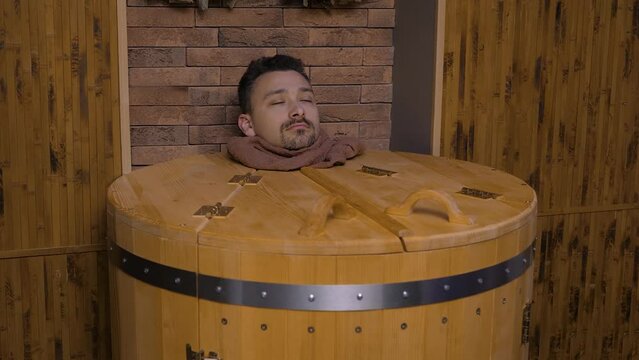 Male head peeks out of cedar hot herbal barrel at spa. Handsome middle-aged man receives sauna procedure with healing effect and enjoys the process of phyto barrel