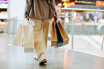 Low section of unrecognizable woman wearing pants and holding blank shopping bags while walking in mall, copy space - obrazy, fototapety, plakaty