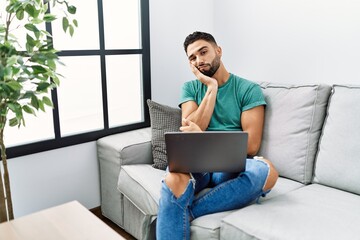 Young handsome man with beard using computer laptop sitting on the sofa at home thinking looking...