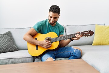 Young arab man playing classical guitar sitting on sofa at home