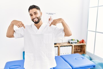 Obraz premium Young handsome man with beard working at pain recovery clinic looking confident with smile on face, pointing oneself with fingers proud and happy.