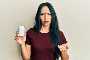 Young hispanic girl holding pills clueless and confused expression. doubt concept.