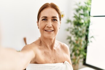 Middle age caucasian woman wearing towel make selfie by the camera sitting on massage board at beauty center