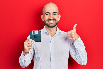 Young bald man holding floppy disk smiling happy and positive, thumb up doing excellent and approval sign