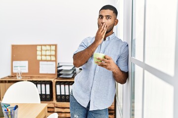 Young indian man drinking a cup coffee at the office covering mouth with hand, shocked and afraid for mistake. surprised expression