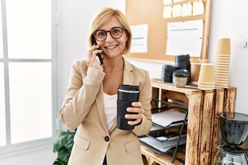 Middle age blonde woman talking on the smartphone drinking coffee at office