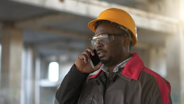 African american worker talks on smartphone at construction site. Builder in yellow hard hat speaks on smartphone at project site