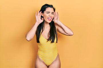 Young hispanic girl wearing swimsuit trying to hear both hands on ear gesture, curious for gossip. hearing problem, deaf