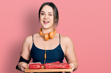 Young hispanic girl wearing workout clothes and holding meat board winking looking at the camera...