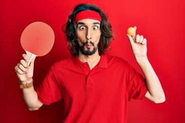 Young hispanic man holding red ping pong racket and ball making fish face with mouth and squinting...