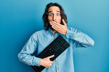 Young hispanic man holding keyboard covering mouth with hand, shocked and afraid for mistake....