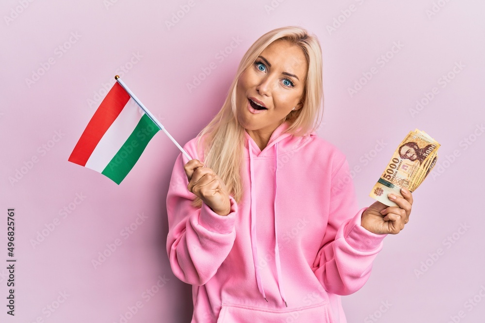 Wall mural young blonde woman holding hungary flag and forint banknotes afraid and shocked with surprise and am