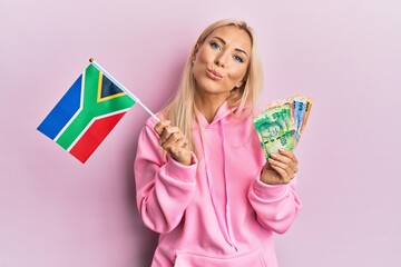 Young blonde woman holding south africa flag and rand banknotes looking at the camera blowing a kiss being lovely and sexy. love expression.