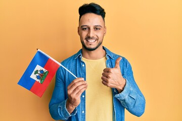 Young arab man holding haiti flag smiling happy and positive, thumb up doing excellent and approval sign