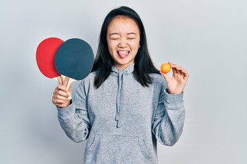 Young chinese girl holding red ping pong rackets and ball sticking tongue out happy with funny...