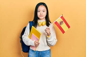 Young chinese girl exchange student holding spanish flag relaxed with serious expression on face. simple and natural looking at the camera.