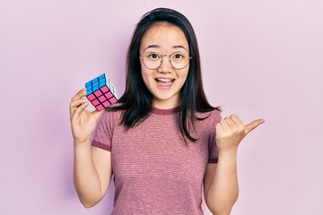Young chinese girl playing colorful puzzle cube intelligence game pointing thumb up to the side smiling happy with open mouth