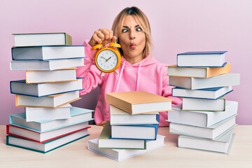 Young caucasian woman holding alarm clock sitting on the table with books making fish face with mouth and squinting eyes, crazy and comical.