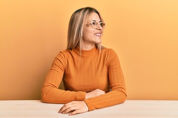 Young caucasian woman wearing casual clothes and glasses sitting on the table looking away to side with smile on face, natural expression. laughing confident.