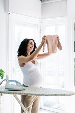 pregnant woman by the bright windows of her house ironing and folding the clothes of her future baby. organization and preparation of the room of the newborn