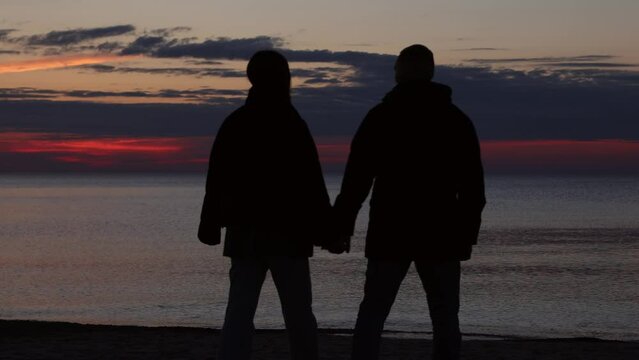 young couple silhouettes walking in sunset on beach, two lovers holding hands walking towards sea ocean in dark evening, relations, love emotions, date, sweet memories after vacations 
