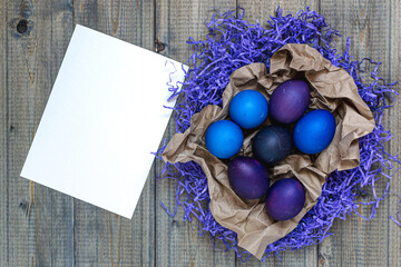 Flat lay top view of a rustic style wooden table on which dyed Easter eggs in very peritoneal and purple with blue lie on paper filler and brown craft paper, next to it lies a sheet of paper, mockup.