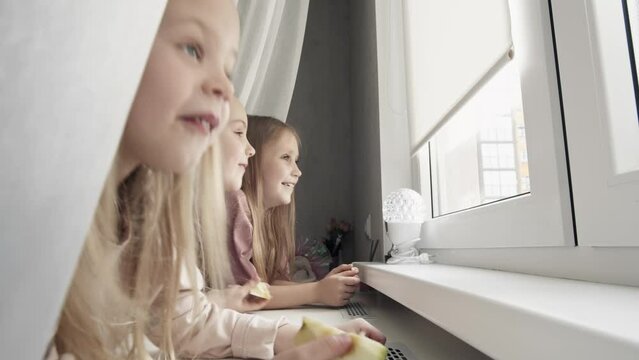 Close-up of three little girls, siblings watching out of window, looking at the street and laughing. Sisters spending time together at home. Family togetherness concept.